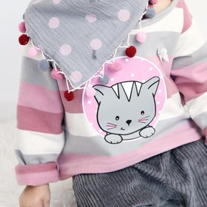 Pode incluir: Um conjunto infantil com uma camisa de mangas compridas &agrave;s riscas cinza e rosa com um gr&aacute;fico de gato. O conjunto inclui cal&ccedil;as de veludo cinzentas e um cachecol cinzento com bolinhas, adornado com pompons. O esquema de cores &eacute; suave e pastel.