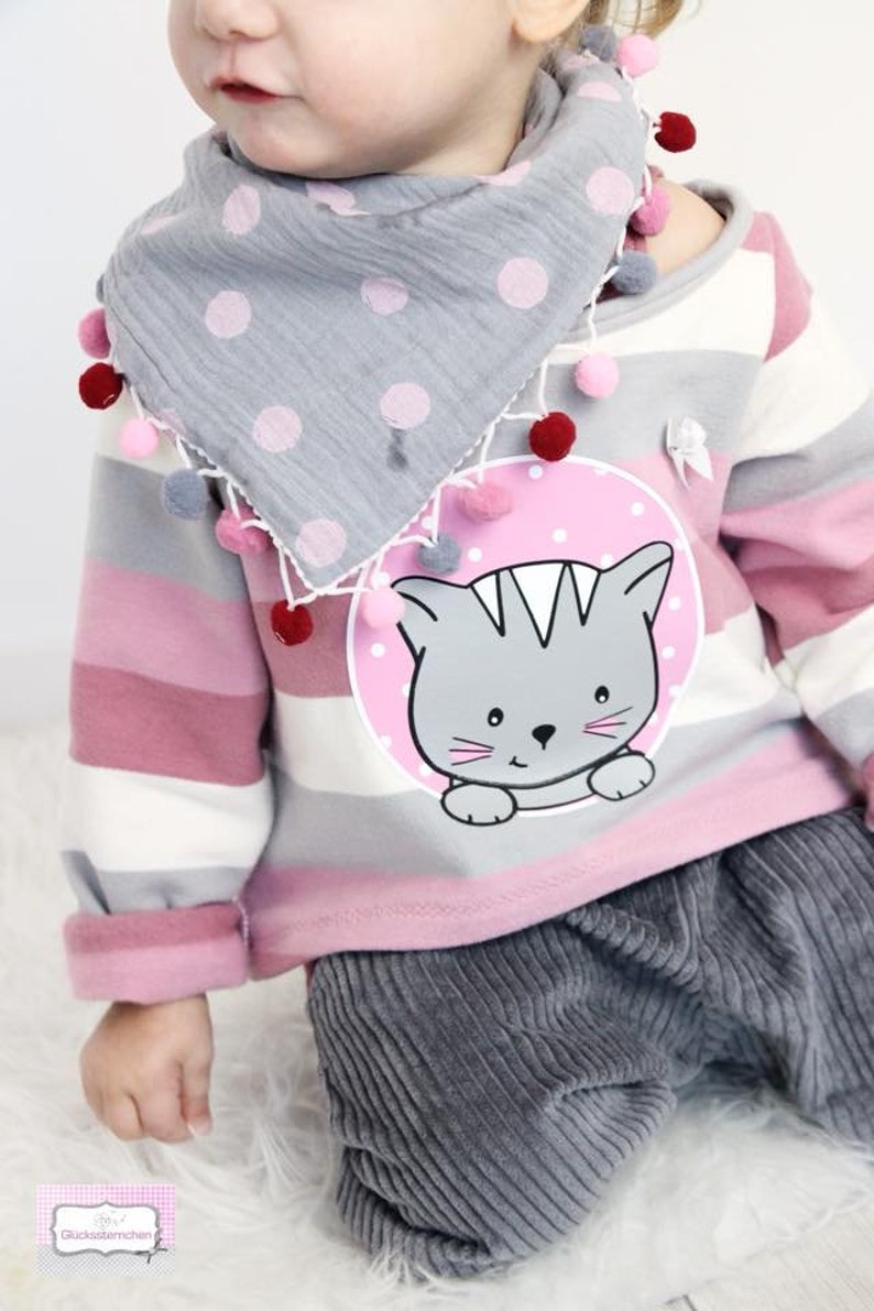 Pode incluir: Um conjunto infantil com uma camisa de mangas compridas &agrave;s riscas cinza e rosa com um gr&aacute;fico de gato. O conjunto inclui cal&ccedil;as de veludo cinzentas e um cachecol cinzento com bolinhas, adornado com pompons. O esquema de cores &eacute; suave e pastel.
