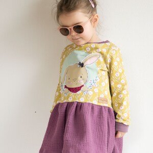 Op de afbeelding: Een jong meisje draagt een gele en groene jurk met een konijnenapplicatie. De jurk heeft een paarse rok met een gerimpelde zoom. Het meisje draagt een roze zonnebril en heeft haar haar in een knotje.