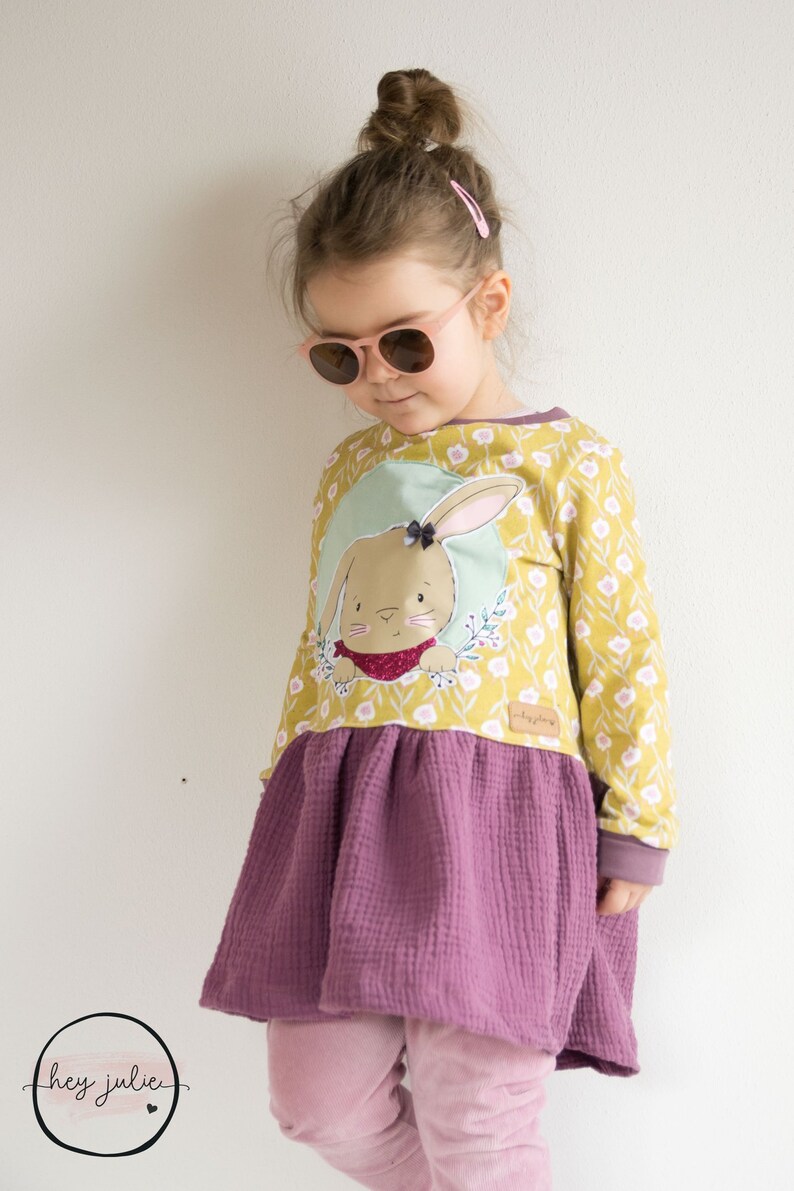 Op de afbeelding: Een jong meisje draagt een gele en groene jurk met een konijnenapplicatie. De jurk heeft een paarse rok met een gerimpelde zoom. Het meisje draagt een roze zonnebril en heeft haar haar in een knotje.