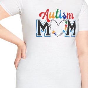 Peut inclure: T-shirt blanc avec l'inscription "Autism Mom" dans un design coloré. Le mot "Autism" est en rouge et en couleurs arc-en-ciel, avec les lettres "MOM" en noir et bleu. Une forme de cœur est remplie de pièces de puzzle.