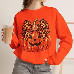 May include: A bright orange long-sleeve sweatshirt featuring a large graphic of a glittery orange jack-o'-lantern pumpkin adorned with a prominent leopard print bow. The pumpkin design includes a classic carved face with triangular eyes and a grin.