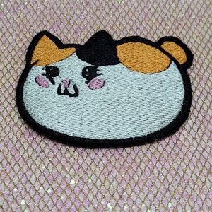 May include: A white, orange, and black embroidered patch of a cartoon cat with pink cheeks. The cat is round and has a simple, friendly expression.