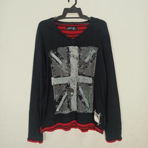 Könnte beinhalten: Schwarzes Langarmshirt mit einem verblichenen Union Jack-Motiv. Das V-Ausschnitt hat ein rot-schwarz gestreiftes Futter, und die Manschetten und der Saum sind mit roten Streifen versehen. Ein kleines Etikett ist unten rechts sichtbar.