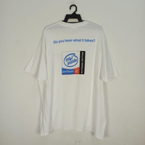 Puede incluir: Camiseta blanca con un logotipo de Intel Inside Pentium 4 Extreme Edition azul y rojo en la parte posterior. La camiseta tiene el texto "Do you have what it takes?" impreso sobre el logotipo.