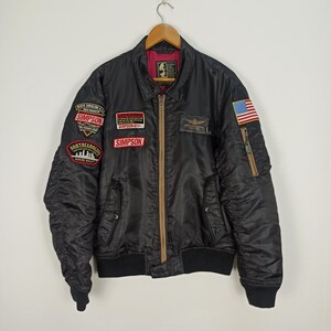 Op de afbeelding: Zwarte bomberjack met meerdere patches, waaronder een Amerikaanse vlag en de naam "SIMPSON". De jas heeft een bruine rits en geribde manchetten en zoom. De jas hangt aan een houten kleerhanger.