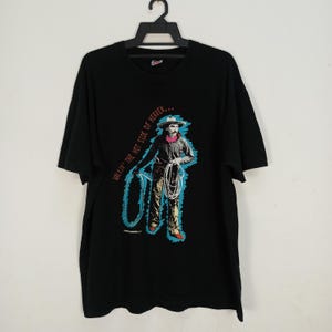 Op de afbeelding: Zwart T-shirt met een afbeelding van een cowboy met een lasso en een touw. De cowboy draagt een hoed en een roze sjaal. De tekst "WALKIN' THE HOT SIDE OF HEAVEN..." staat op het shirt.