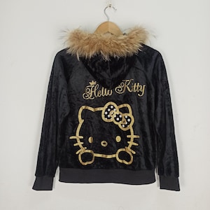 May include: Black velvet zip-up hoodie with a faux fur-trimmed hood. The back features a gold glitter Hello Kitty design. The hoodie is hanging on a wooden hanger.