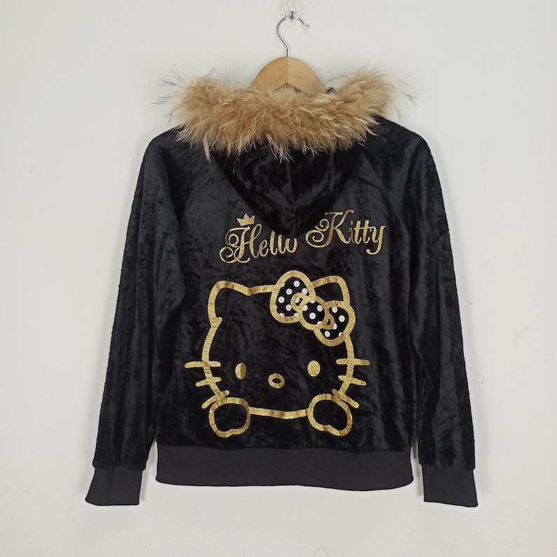 May include: Black velvet zip-up hoodie with a faux fur-trimmed hood. The back features a gold glitter Hello Kitty design. The hoodie is hanging on a wooden hanger.