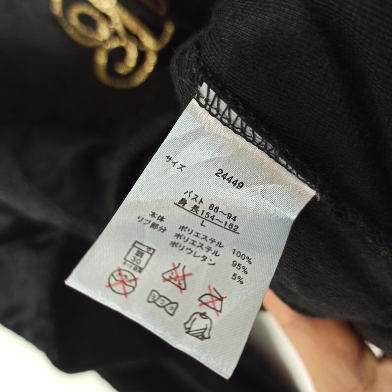 May include: Black garment tag with Japanese text and care symbols. The tag includes size information, measurements in centimeters, and fabric composition details. The main fabric is 100% polyester, with a rib section of 95% polyester and 5% polyurethane.