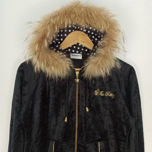 May include: Black hooded jacket with a faux fur trim around the hood. The jacket features a gold zipper and the words "Hello Kitty" embroidered in gold. The hood lining has a white polka dot pattern on a black background.