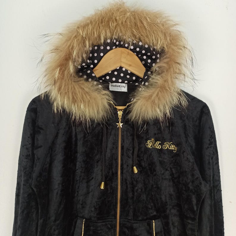 May include: Black hooded jacket with a faux fur trim around the hood. The jacket features a gold zipper and the words "Hello Kitty" embroidered in gold. The hood lining has a white polka dot pattern on a black background.