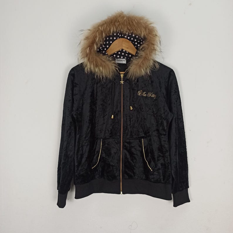 May include: Black velvet zip-up hoodie with a faux fur-trimmed hood. The hood lining features a white polka dot pattern. The hoodie has a gold zipper and the words "Hello Kitty" embroidered in gold on the chest.