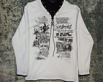 Pode incluir: Uma camisa branca de manga comprida com acabamento preto nos punhos e gola interna preta. A frente apresenta um decote com cordões e ilhós pretos, além de extensos textos e gráficos de caveiras impressos, incluindo a palavra Detroit.