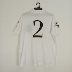 May include: White polo shirt with a black number "2" on the back and a small embroidered patch on the right sleeve that says "POLO RALPH LAUREN" and features a Scottish flag.