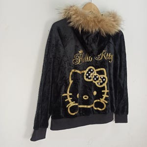 May include: Black hooded jacket with a faux fur trim around the hood. The back features a gold glitter Hello Kitty design with the words "Hello Kitty" in gold script. The jacket has a black zipper and black cuffs.