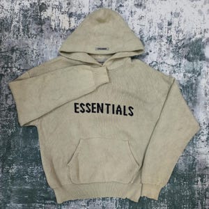 Op de afbeelding: Beige gebreide hoodie met het woord "ESSENTIALS" in zwarte letters op de borst. De hoodie heeft een kangoeroezak en een capuchon. De merknaam staat ook op de capuchon. Het kledingstuk is gemaakt van zacht, getextureerd breisel.