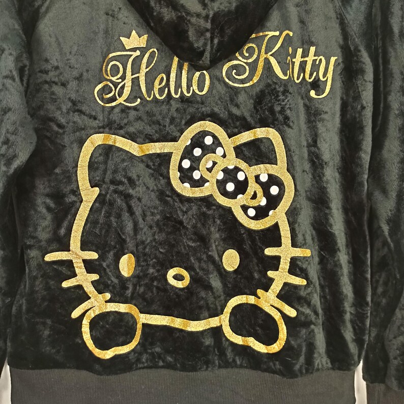 May include: Black hooded sweatshirt featuring a gold Hello Kitty design. The design includes the Hello Kitty logo and a large gold outline of Hello Kitty's face and bow. The sweatshirt is made of a soft, velvet-like material.