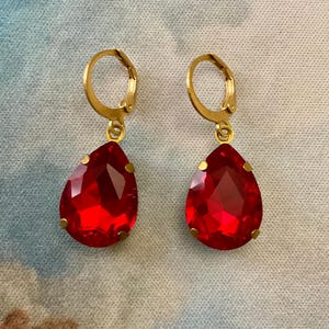 Peut inclure: Une paire de boucles d'oreilles à fermoir à levier dorées avec de grandes pierres précieuses rouges en forme de goutte d'eau. Les boucles d'oreilles sont présentées sur un fond à motifs bleu clair et blanc.