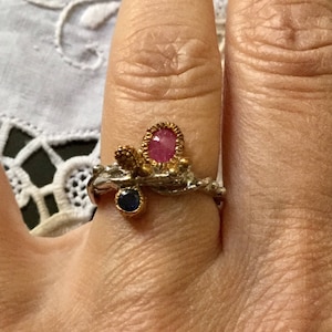 Peut inclure: Une bague en argent avec une bande en or et deux pierres précieuses. La plus grande pierre est rose et de forme ovale, tandis que la plus petite est bleue et ronde. La bague est sertie dans un design torsadé et organique.