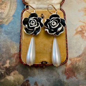 Puede incluir: Un par de pendientes elegantes con la parte superior en forma de rosa en blanco y negro y colgantes en forma de lágrima nacarada. Los pendientes se muestran dentro de un joyero vintage con interior dorado.