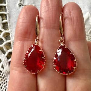 Peut inclure: Une paire de boucles d'oreilles en forme de goutte rouge avec des montures dorées. Les boucles d'oreilles ont un design facetté et pendent d'un crochet.