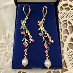 Peut inclure: Paire de boucles d'oreilles pendantes dorées avec un motif floral, des détails verts et roses. Les boucles d'oreilles ont une structure en forme de branche avec des pierres roses et une perle blanche en bas. Présentées dans un écrin bleu. Mesurent environ 8 cm de long.