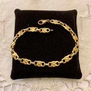 Peut inclure: Un bracelet doré avec un motif de chaîne tressée, présenté sur une surface de velours noir. Le bracelet est doté d'une fermeture à fermoir mousqueton. Le bijou est présenté sur un napperon en dentelle blanche.