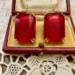 Peut inclure: Une paire de boucles d'oreilles en pierres précieuses rouges avec des montures dorées. Les boucles d'oreilles sont dans une petite boîte rouge.