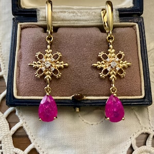 Peut inclure: Boucles d'oreilles dorées avec un motif de flocon de neige orné de petits diamants et une pierre précieuse rose vif en forme de goutte. Les boucles d'oreilles sont présentées dans un écrin à bijoux bleu vintage.