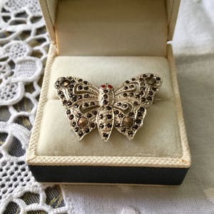 Peut inclure: Une broche en forme de papillon argentée avec des strass noirs et rouges. La broche est dans une boîte blanche avec une doublure bleue.