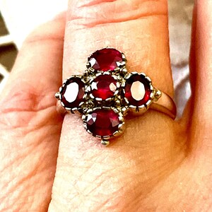 Peut inclure: Bague argentée ornée de cinq pierres précieuses ovales rouge foncé, disposées en grappe. Les pierres sont serties individuellement, créant un motif floral ou en forme de croix. La bague est portée sur un doigt.