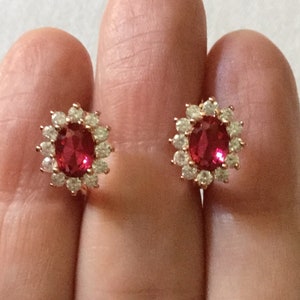 Peut inclure: Une paire de boucles d'oreilles à clous avec une pierre précieuse rouge centrale de taille ovale, entourée d'un halo de douze petites pierres blanches rondes et transparentes, le tout monté sur une structure en métal doré aux tons chauds.