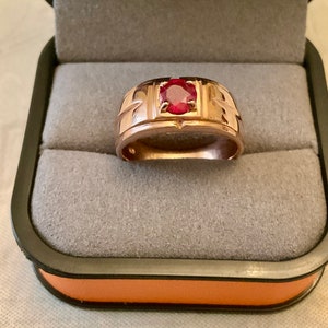 Peut inclure: Une bague en or avec une pierre précieuse rouge sertie dans un bézel carré. La bague a une large bande avec un motif décoratif.
