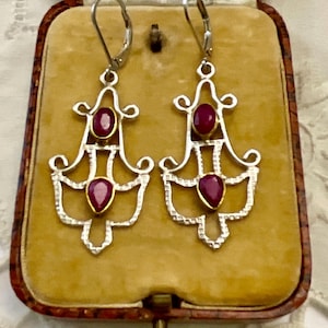 Può includere: Un paio di orecchini a lampadario color argento con un design a filigrana, ciascuno con due pietre preziose ovali rosso scuro. Gli orecchini sono esposti in un portagioie vintage foderato in velluto con esterno marrone.