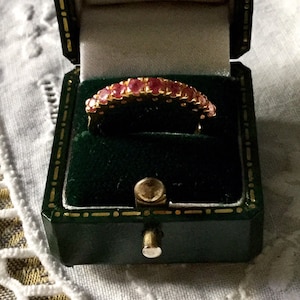 May include: A gold ring with a row of pink gemstones set in a band. The ring is in a green velvet ring box.