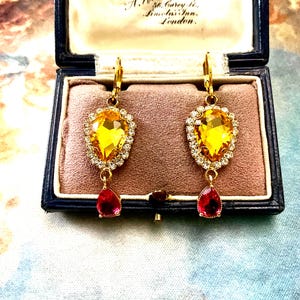 Peut inclure: Paire de boucles d'oreilles pendantes dorées avec de grandes pierres précieuses jaunes en forme de goutte, entourées de petits cristaux transparents. Chaque boucle d'oreille a une petite pierre rouge en forme de goutte. Présentées dans un écrin à bijoux bleu vintage.