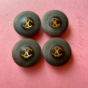 Peut inclure: Quatre boutons gris foncé avec un motif double-C doré au centre. Les boutons sont ronds et ont un centre légèrement surélevé. Le fond est de couleur rose clair.