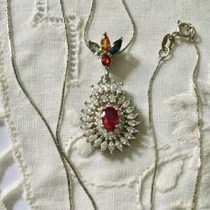Peut inclure: Un collier en argent avec un pendentif composé d'un gros rubis rouge entouré de petits diamants blancs. Le pendentif comporte également un groupe de petites pierres précieuses colorées au centre.