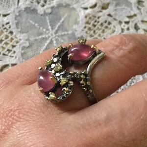 Peut inclure: Une bague argentée ornée de trois pierres précieuses roses ovales. La bague présente un design organique unique avec de petits accents jaune-vert. La bague est portée sur un doigt, mettant en valeur ses détails et sa palette de couleurs.