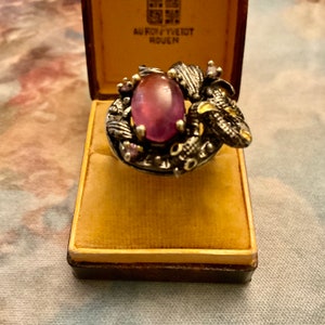 Peut inclure: Une bague en argent avec une grande pierre précieuse ovale de couleur violette sertie dans un design texturé détaillé. La bague est dans une petite boîte brune avec le texte "AU ROY YVE TOT ROUEN" sur le couvercle.