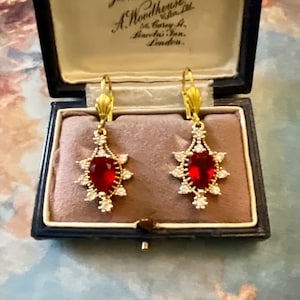 Peut inclure: Une paire de boucles d'oreilles pendantes en or avec des pierres rouges en forme de poire entourées de petites pierres claires. Les boucles d'oreilles sont dans une boîte en velours bleu avec une étiquette qui indique "A. Woodhouse Ltd. 56, Carey St. Lincoln's Inn, London."