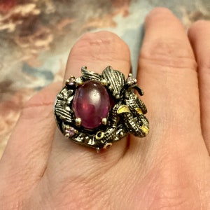 Peut inclure: Une bague en argent avec une grande pierre précieuse ovale de couleur violette entourée de petites pierres précieuses roses. La bague présente un design détaillé d'un serpent avec des accents dorés.