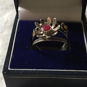 May include: A silver ring featuring a red gemstone at its centre, surrounded by a silver flower design. The ring is displayed on a black velvet background.
