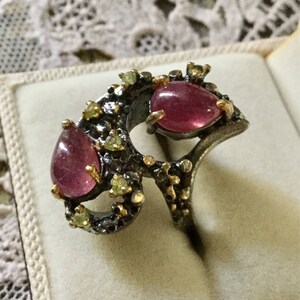 Peut inclure: Une bague en argent avec un design unique mettant en vedette deux pierres précieuses roses et de plus petites pierres précieuses vertes. La bague a une finition texturée et ornée.