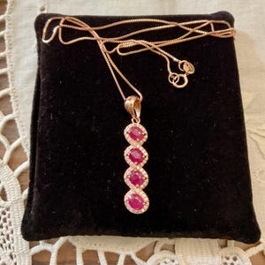 Peut inclure: Un collier en or rose avec un pendentif orné de quatre pierres précieuses rouges ovales entourées de petites pierres claires. Le collier est présenté sur une surface en velours noir.