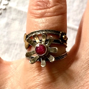 Peut inclure: Une bague argentée avec un motif floral central orné d'une pierre précieuse rouge ronde. La bague a un anneau texturé et des détails dorés. La bague est portée au doigt.