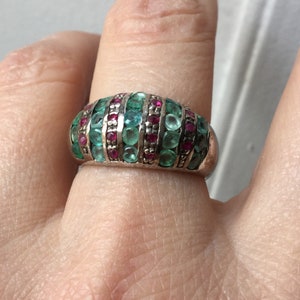 Pode incluir: Um anel de prata com um design abobadado, cravejado com fileiras alternadas de pedras preciosas verde esmeralda e vermelho rubi. O anel é usado em um dedo, mostrando as cores vibrantes e os detalhes intrincados da joia.