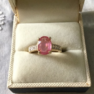 Peut inclure: Une bague en or avec une grande pierre précieuse ovale rose entourée de petites pierres précieuses claires.