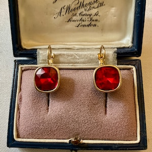 Peut inclure: Boucles d'oreilles dorées avec de grands carrés rouges facettés. Elles sont présentées dans un écrin bleu vintage. Le couvercle de la boîte porte l'inscription "Jewellers Estd. 1692. A. Woodhouse & Son Ltd."